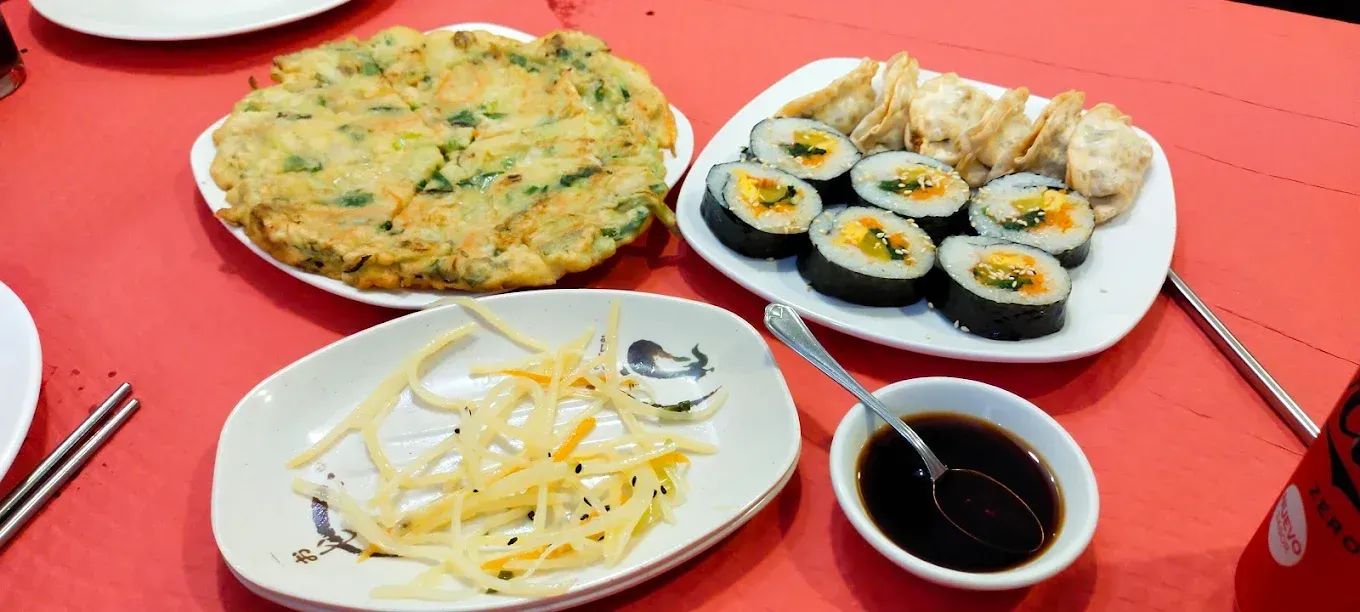 Platos de comida coreana sobre una mesa roja, con tortilla, kimbap, patatas fritas y salsa para mojar.