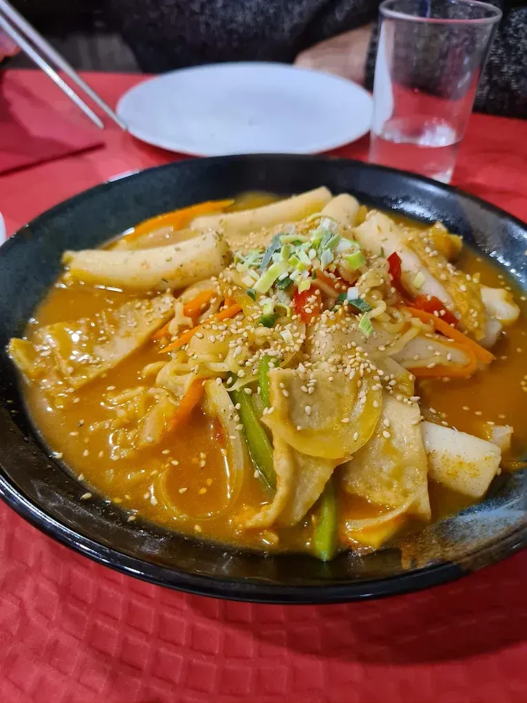 Pasteles de arroz salteados picantes con salsa naranja en una sartén negra, cubiertos con semillas de sésamo y verduras.