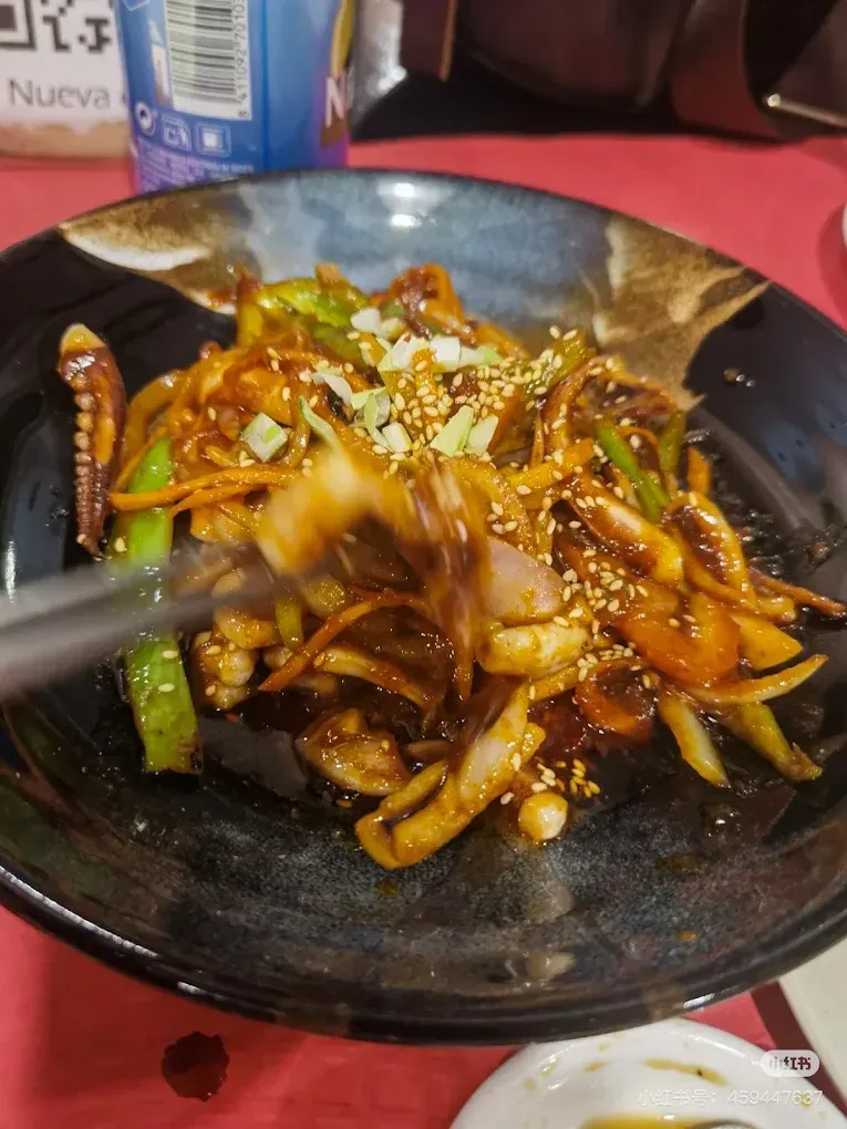 Fideos salteados en un cuenco oscuro con palillos, cubiertos de semillas de sésamo, sobre un mantel rojo.