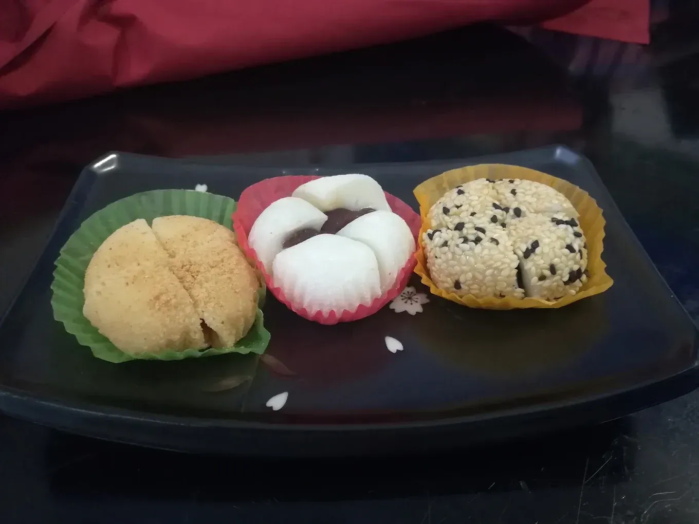 Tres dulces de mochi variados en una bandeja negra: beige, blanco con el borde rojo y cubierto de sésamo.