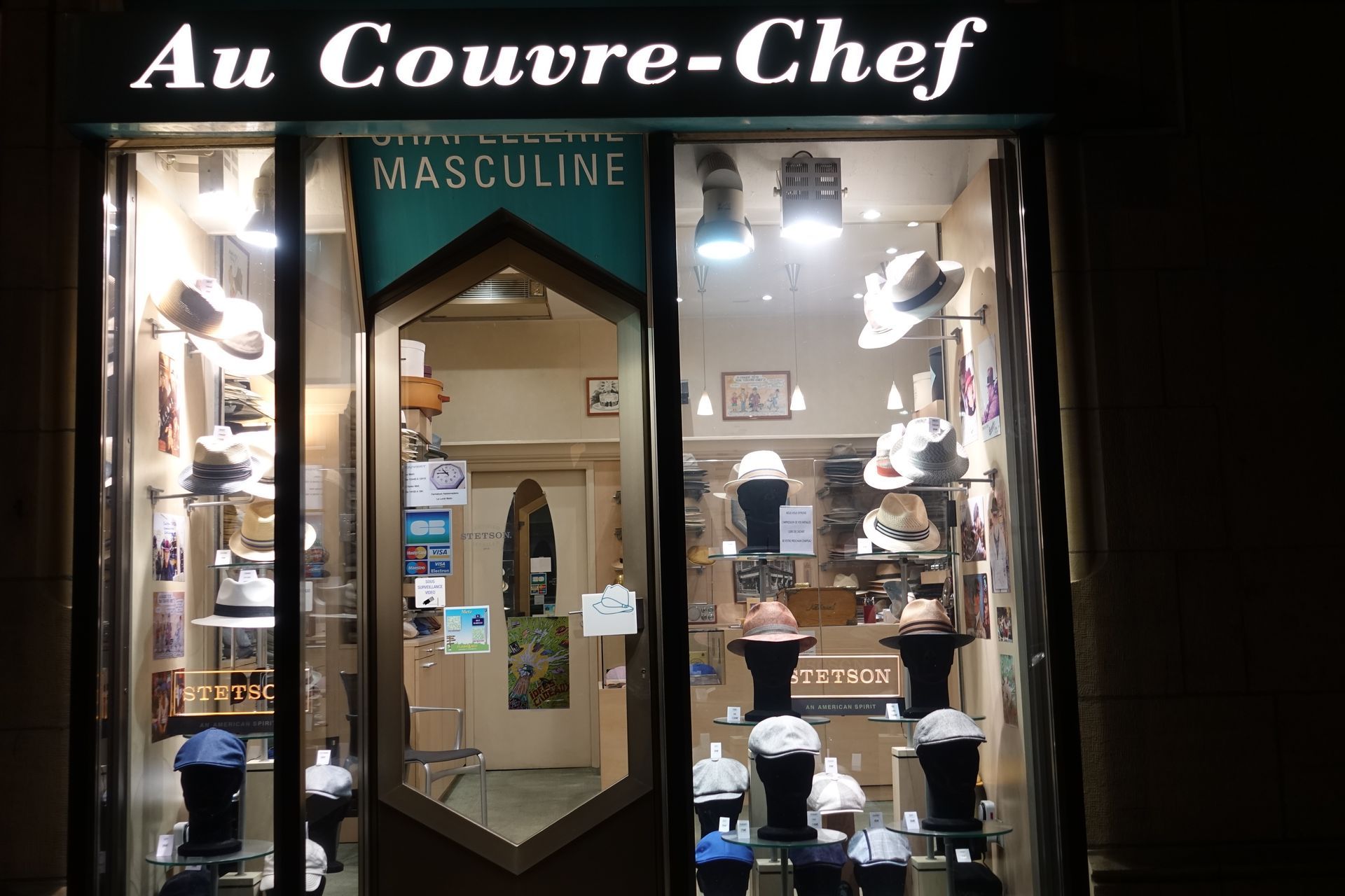 Façade du magasin de chapeaux « Au Couvre-Chef » avec des chapeaux exposés en vitrine la nuit
