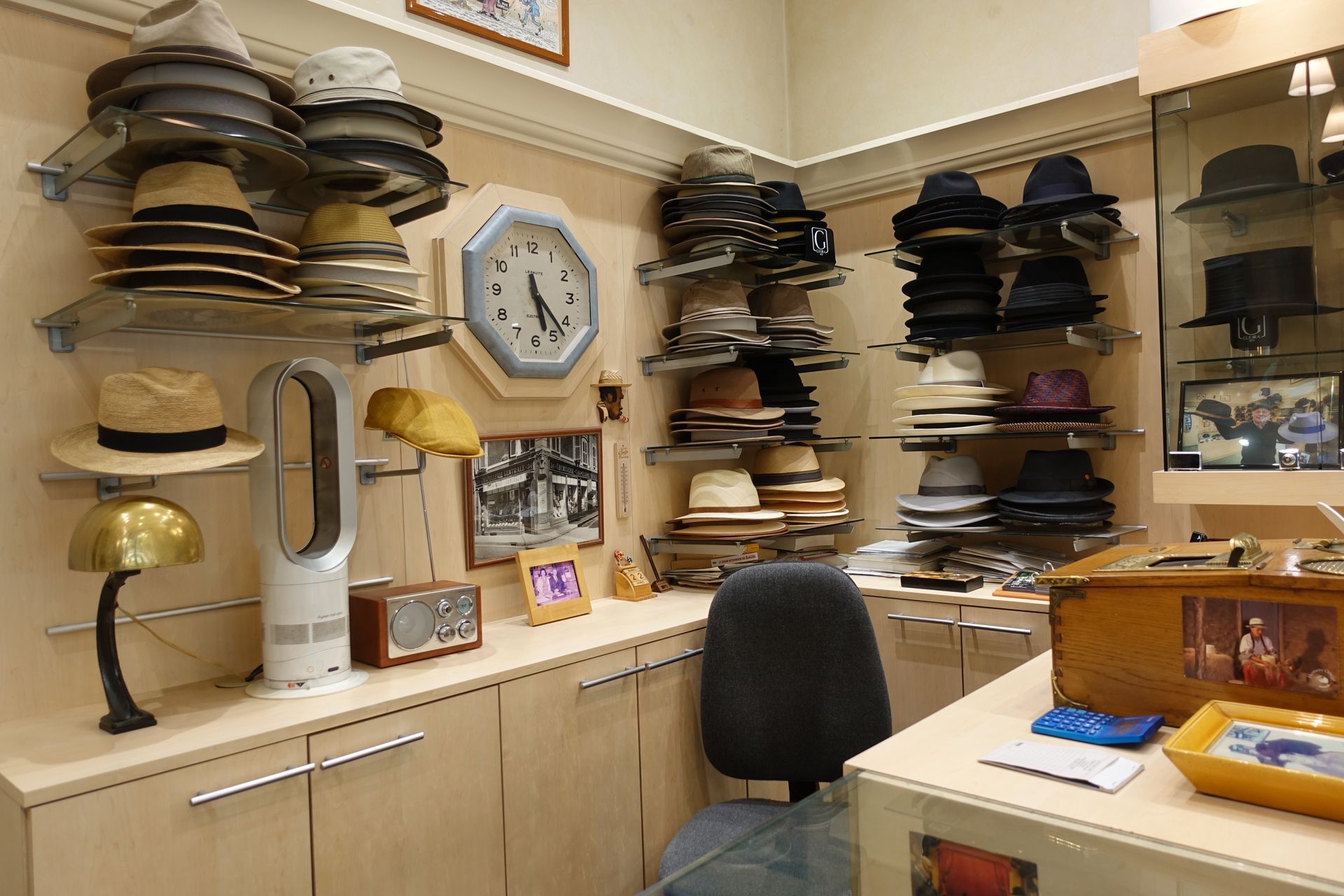 Intérieur d'une petite boutique de chapeaux avec étagères remplies de chapeaux, une horloge et un bureau.