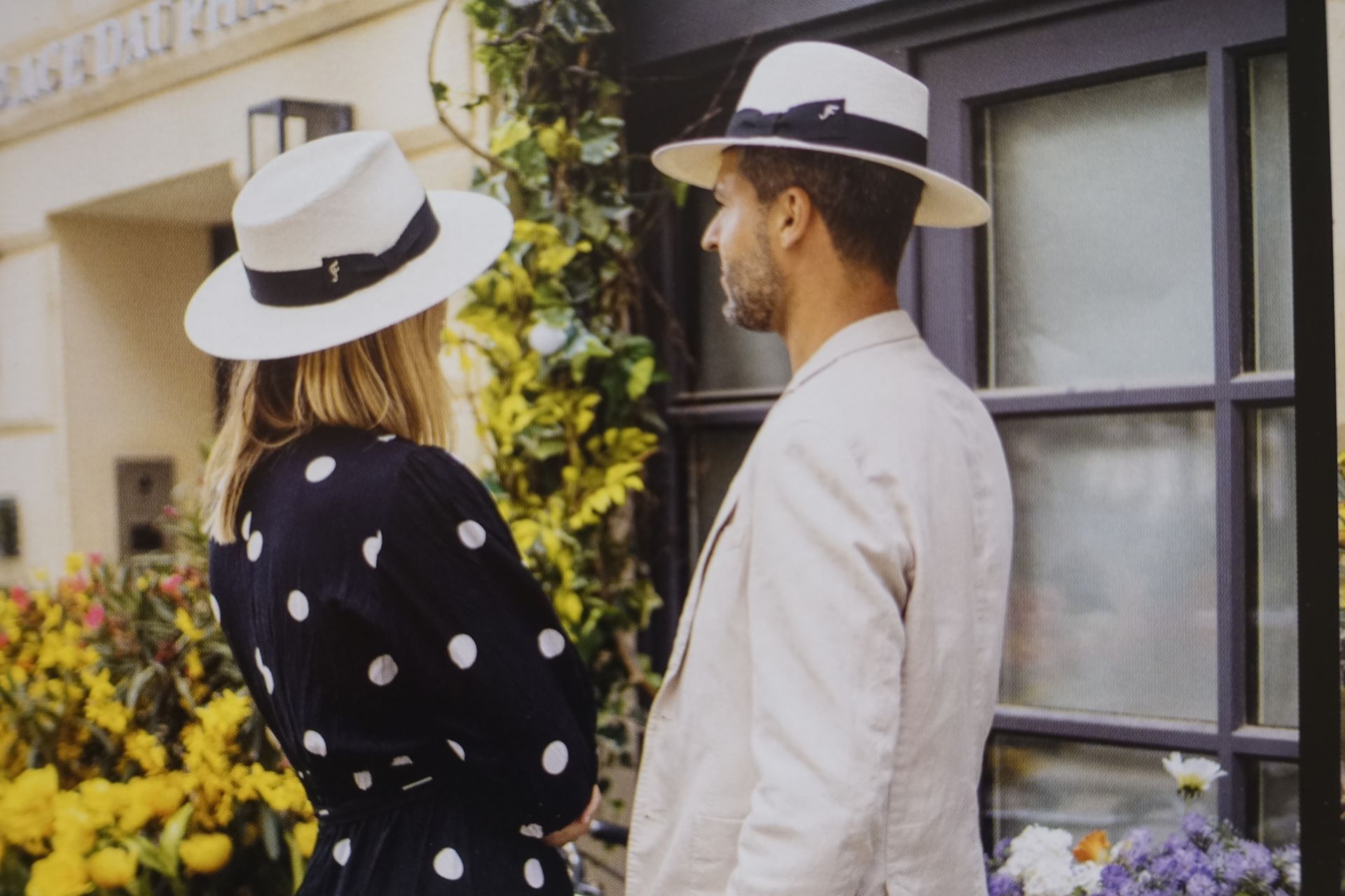 Deux personnes coiffées de chapeaux blancs se tiennent près d'une fenêtre et d'un jardin fleuri.