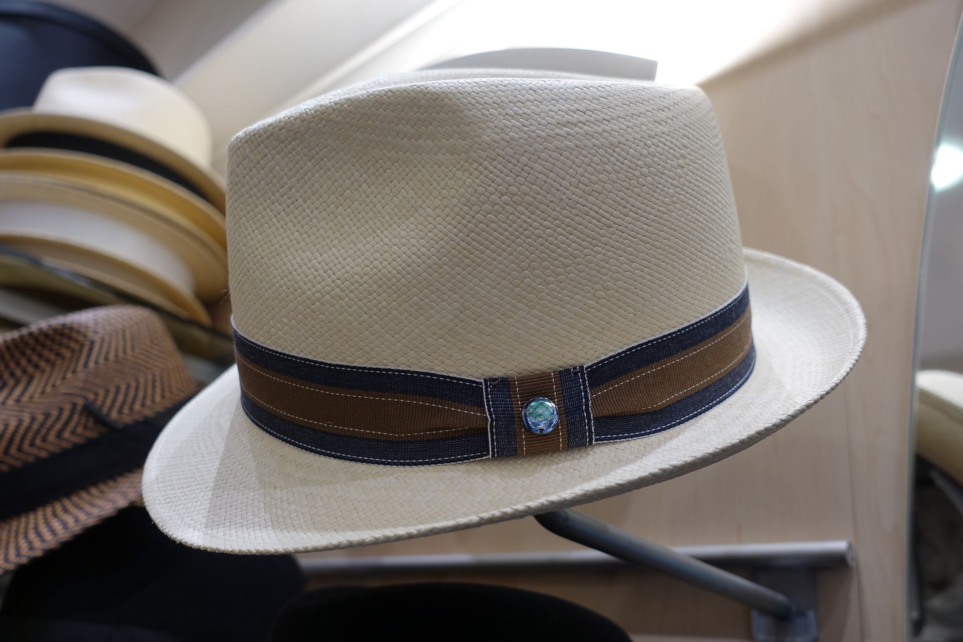 Un chapeau fedora en paille beige avec un ruban marron et une touche de bleu, posé sur une étagère avec d'autres chapeaux.