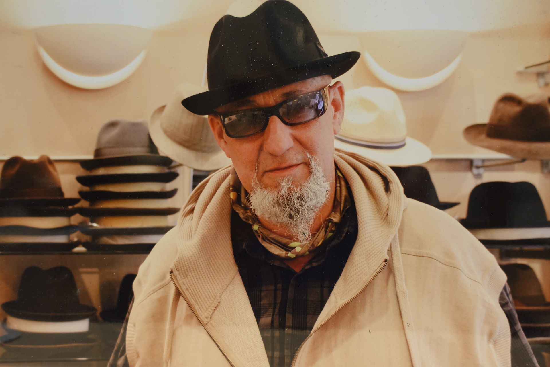 Un homme portant un chapeau noir et des lunettes de soleil dans une chapellerie, avec des étagères de chapeaux derrière lui.