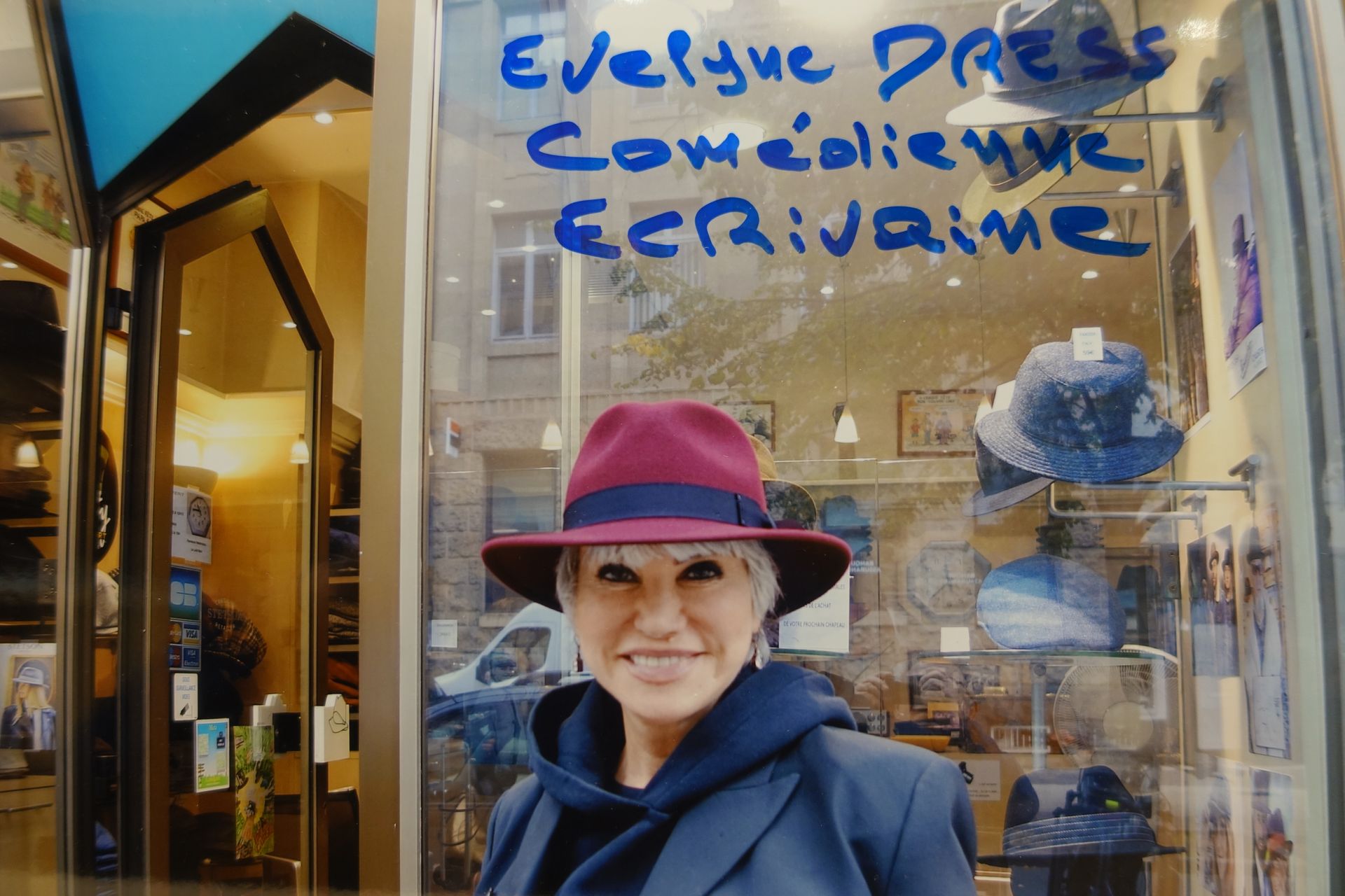 Une femme portant un chapeau rose se tient devant une librairie dont la vitrine est ornée d'une inscription manuscrite bleue.