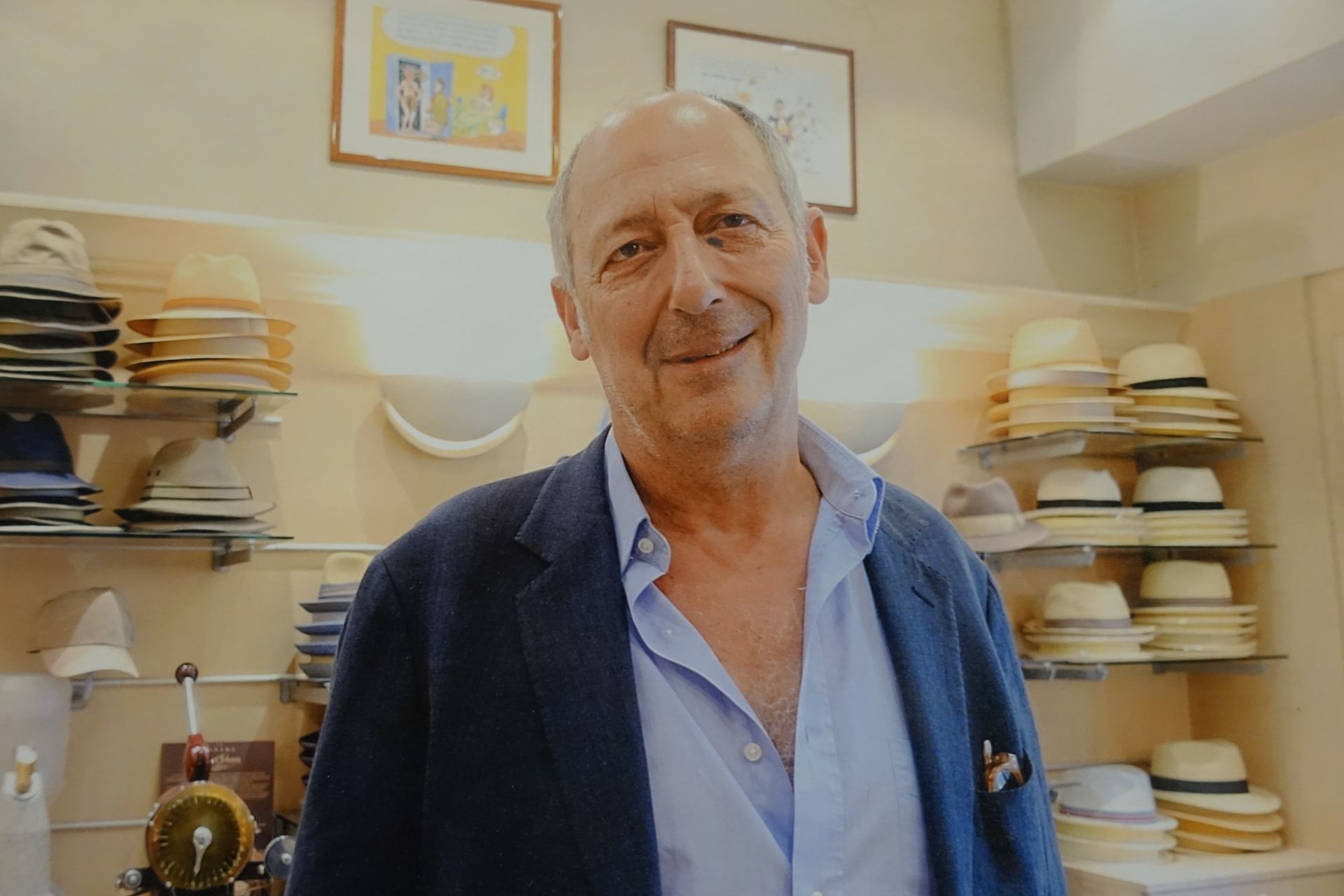 Un homme souriant, vêtu d'un blazer bleu marine, se tient dans une chapellerie, des étagères remplies de chapeaux empilés derrière lui.