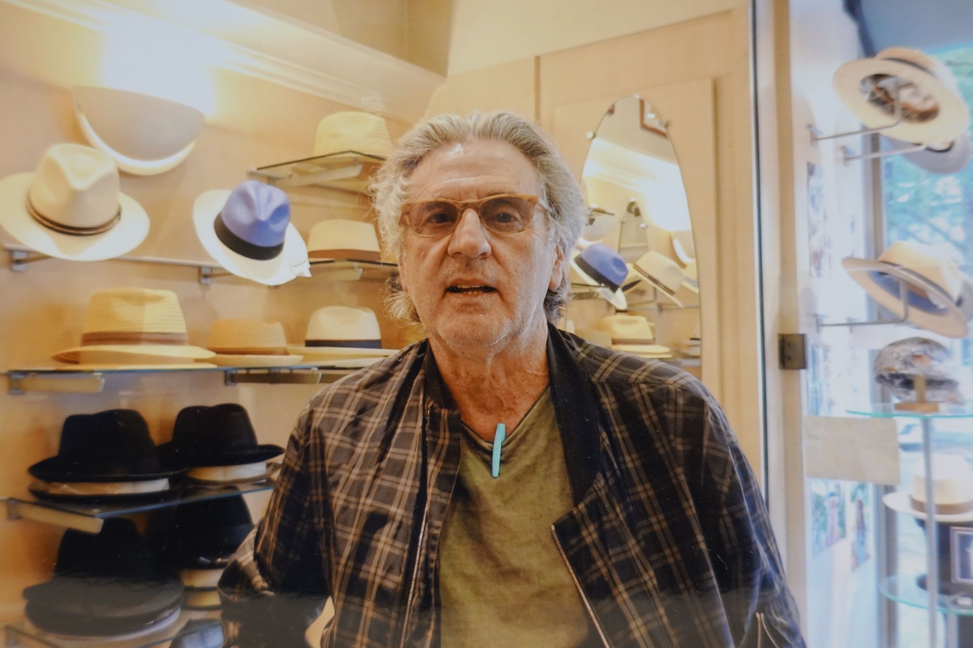 Un homme se tient debout dans une chapellerie, des étagères de chapeaux derrière lui et des lunettes sur le visage.