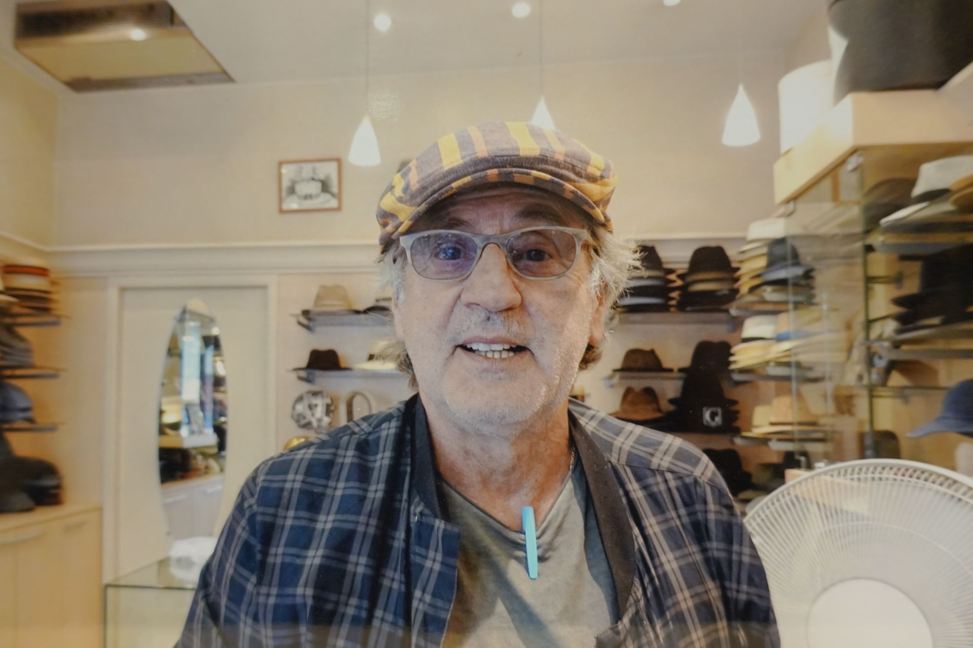 Un homme portant une casquette à carreaux et des lunettes se trouve à l'intérieur d'un magasin lumineux, avec des chapeaux exposés sur des étagères derrière lui.