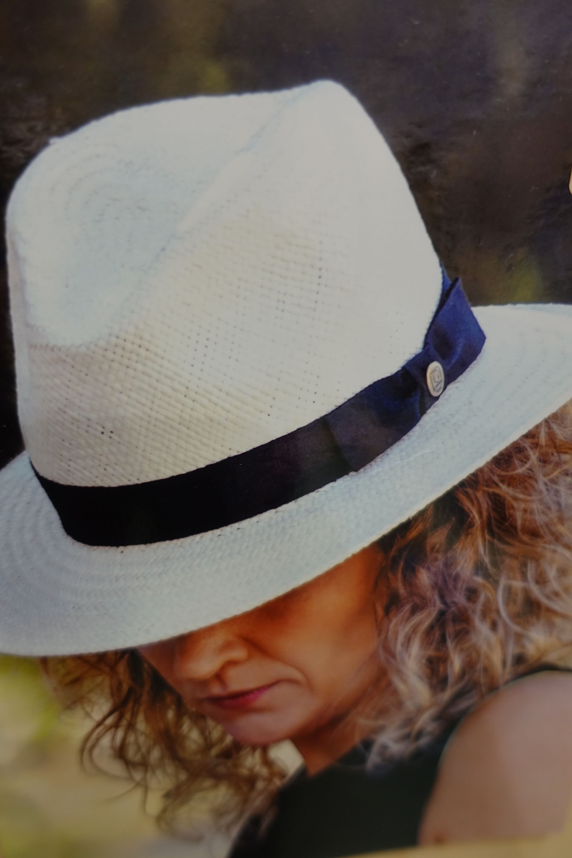 Personne portant un chapeau fedora blanc à ruban noir, regardant vers le bas sur un fond sombre