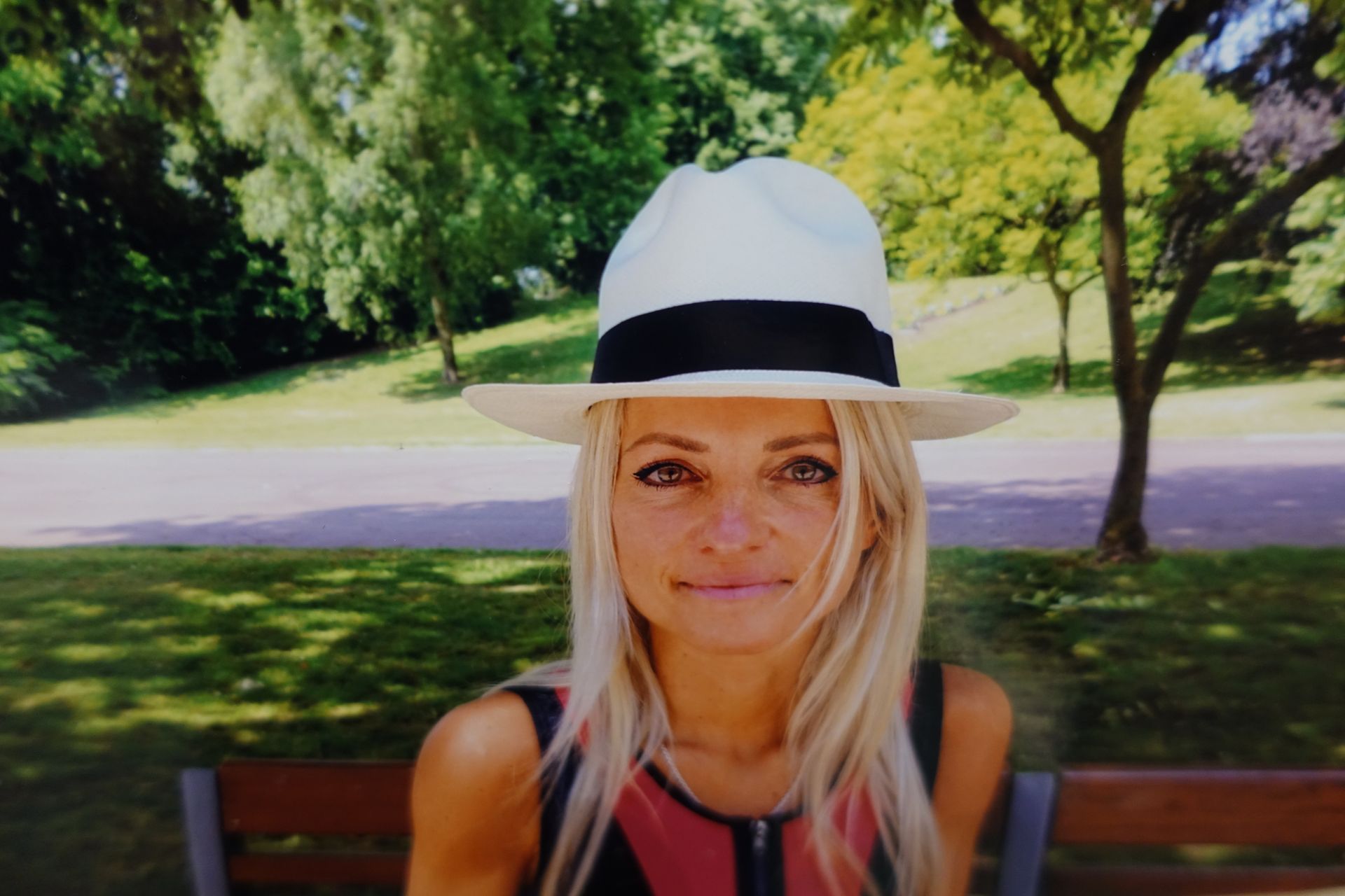 Une femme coiffée d'un chapeau blanc est assise sur un banc dans un parc ensoleillé, bordé d'arbres et d'herbe.