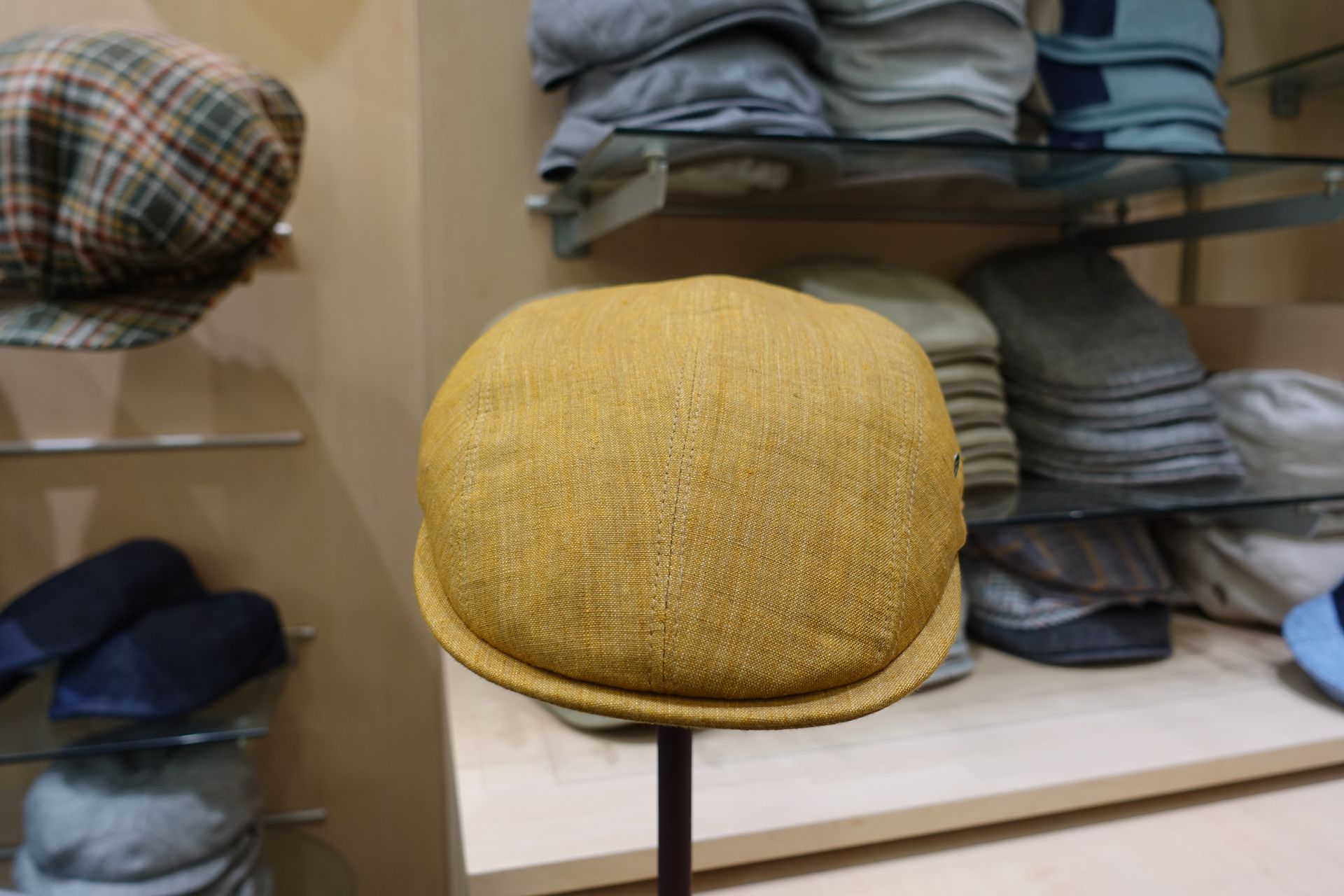 Un bonnet jaune tissé sur un présentoir dans un magasin de vêtements, avec des chapeaux et des pulls pliés sur des étagères derrière.