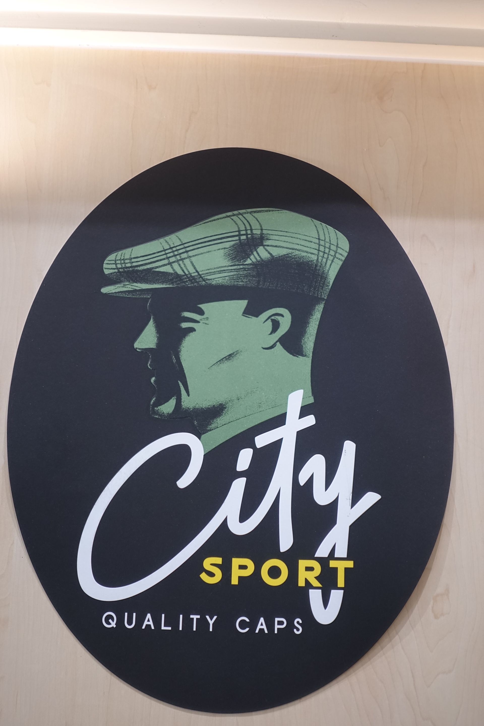 Logo ovale noir « City Sport » avec une illustration de visage vert et le texte « SPORT » en jaune sur un mur clair