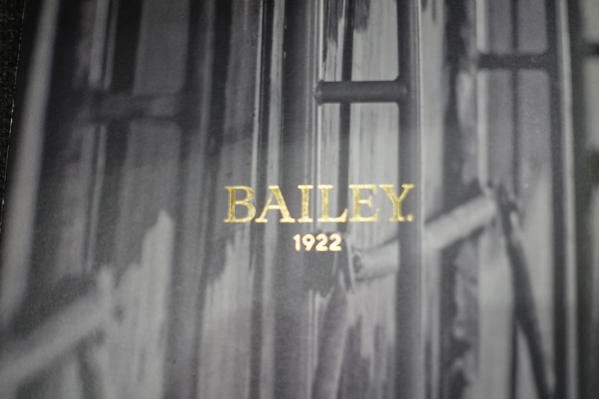 Inscription dorée « BAILEY 1922 » sur un fond noir et blanc flou de barres verticales.