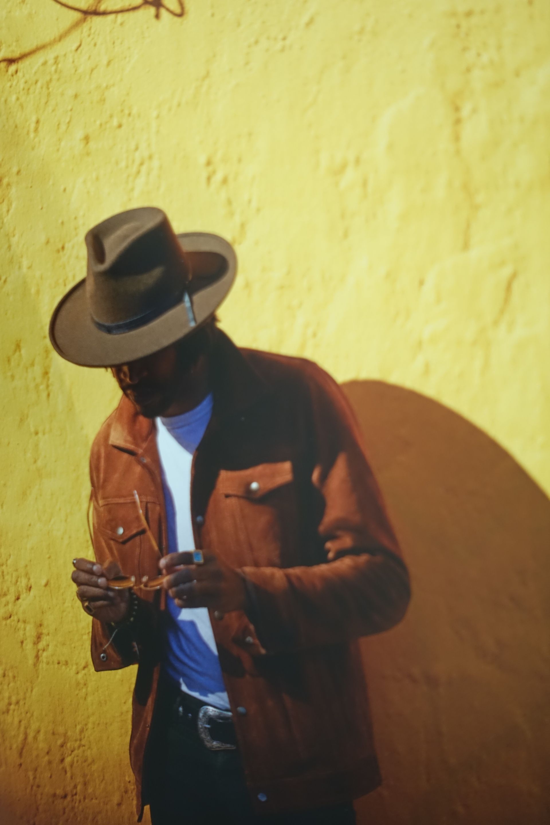 Un homme en veste et chapeau marron se tient près d'un mur jaune, regardant un téléphone.