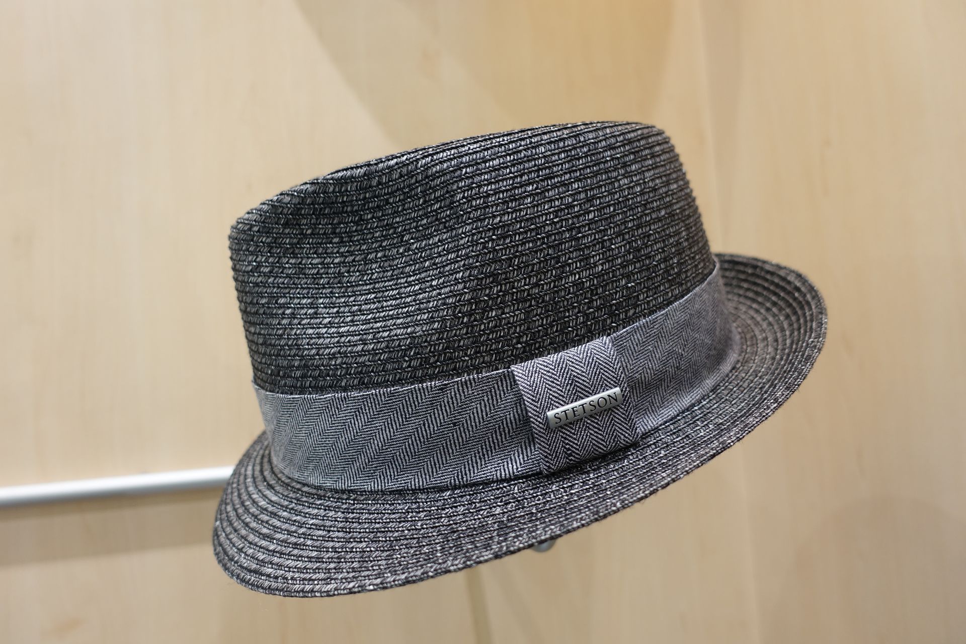 Chapeau fedora noir tissé avec un petit ruban, présenté sur une surface en bois clair.
