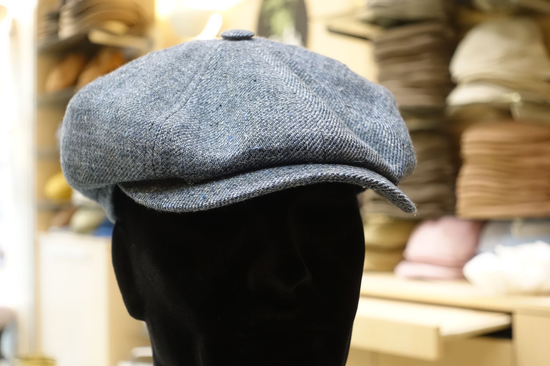 Casquette plate grise sur une tête de mannequin noire dans une chapellerie, avec des étagères de chapeaux en arrière-plan.