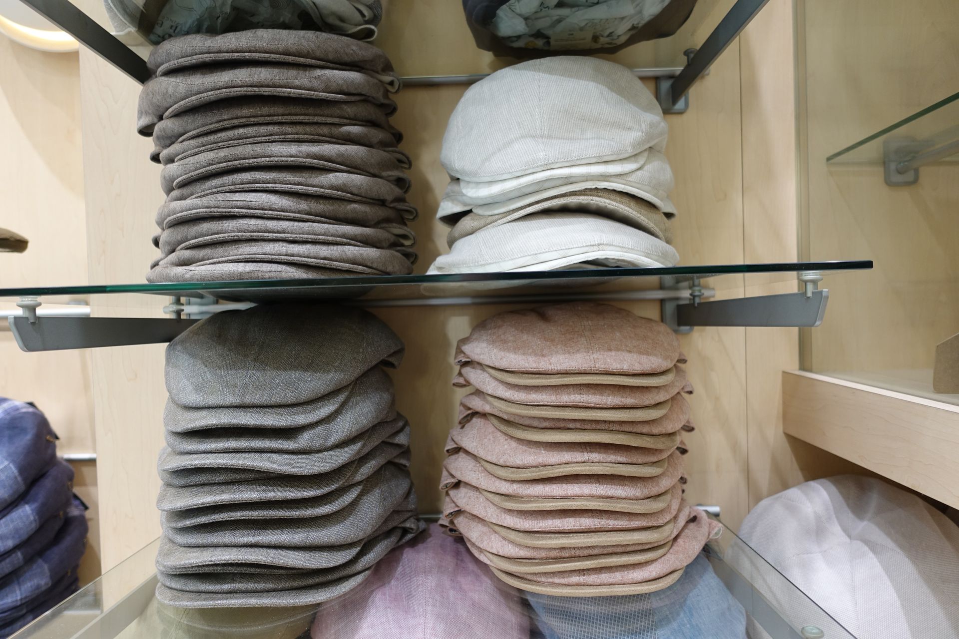 Des piles de bonnets en tricot pliés, de couleurs neutres, sont disposées sur des étagères en verre dans une vitrine de magasin.