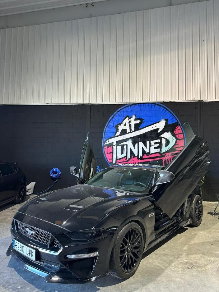 Ford Mustang negro con puertas de ala de gaviota en un garaje, junto a un logotipo 