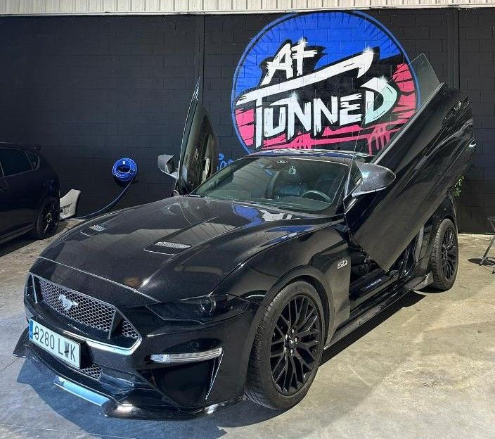 Ford Mustang negro con puertas de ala de gaviota abiertas, estacionado frente a un cartel de 