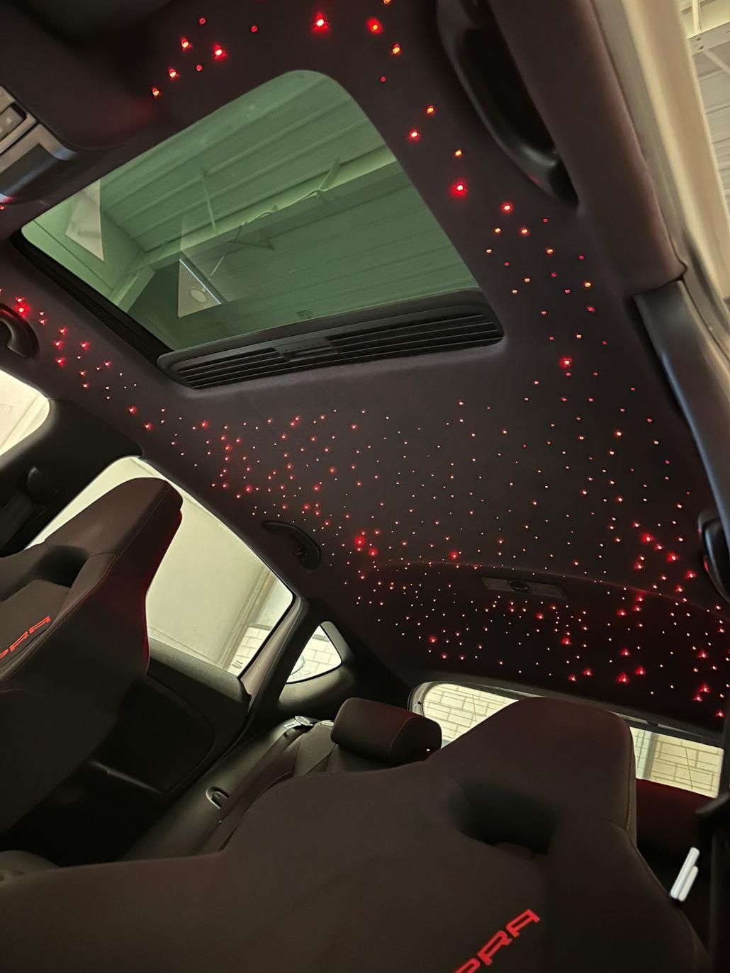 Interior del coche con iluminación de techo tipo cielo estrellado y asientos negros con detalles en rojo.