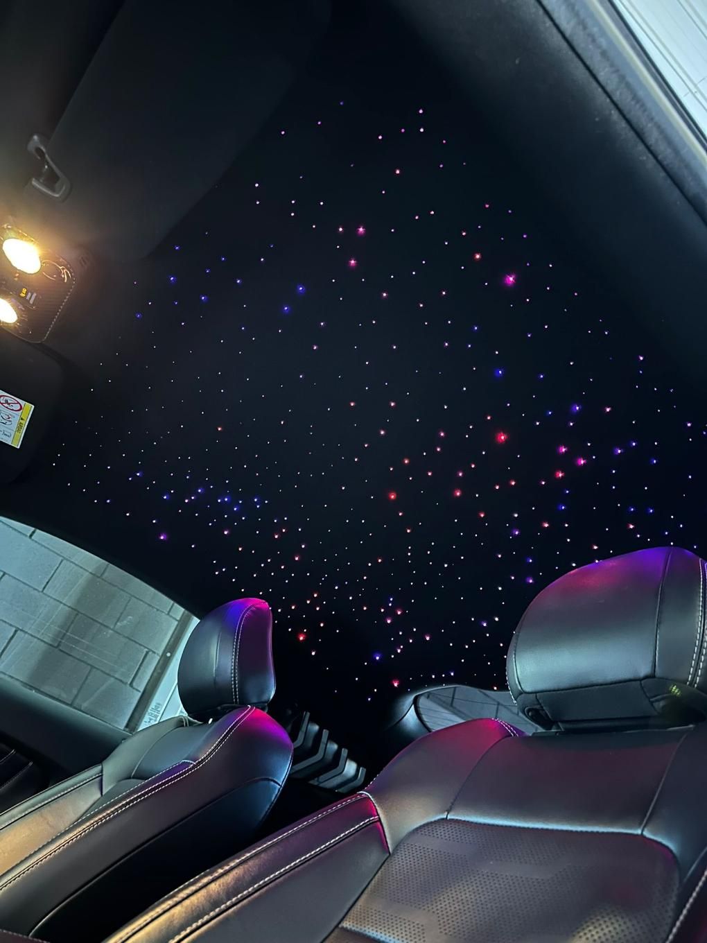 Interior de un automóvil con asientos negros y un techo estrellado iluminado con luces rojas, azules y violetas.