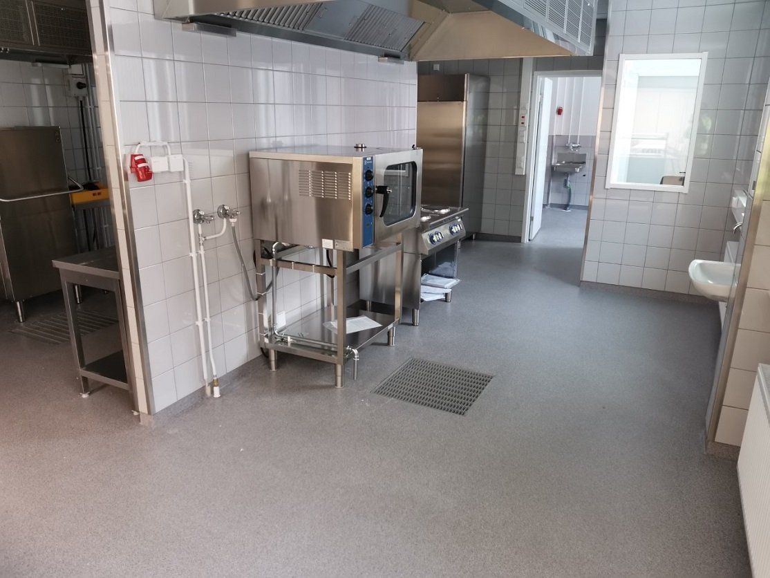 Ammattikeittiön sisustus, jossa ruostumattomasta teräksestä valmistetut kodinkoneet, laatoitetut seinät ja harmaa lattia.