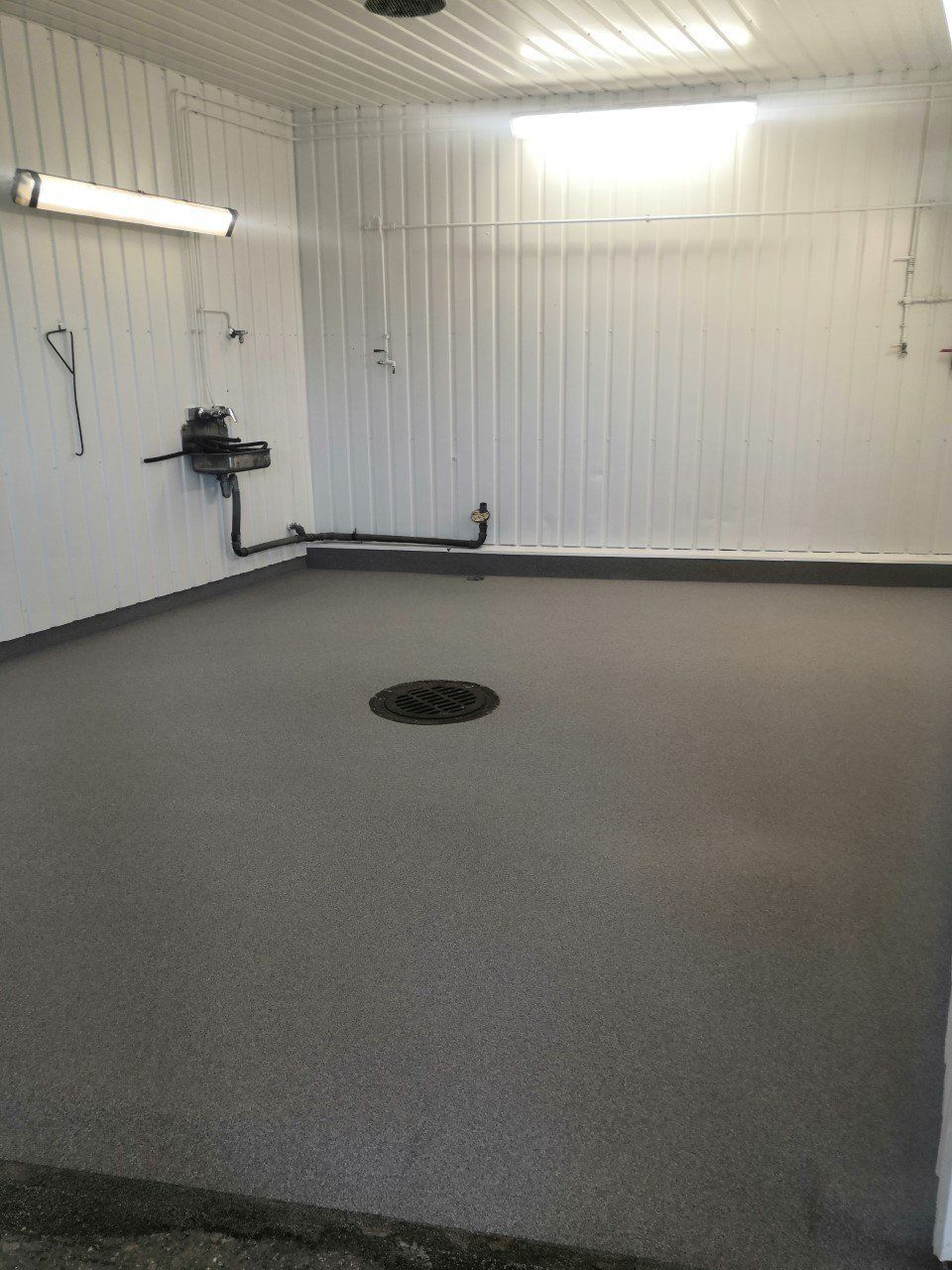 Tyhjä teollisuushuone, jossa on pilkullinen harmaa lattia, valkoiset seinät ja viemäri keskellä.