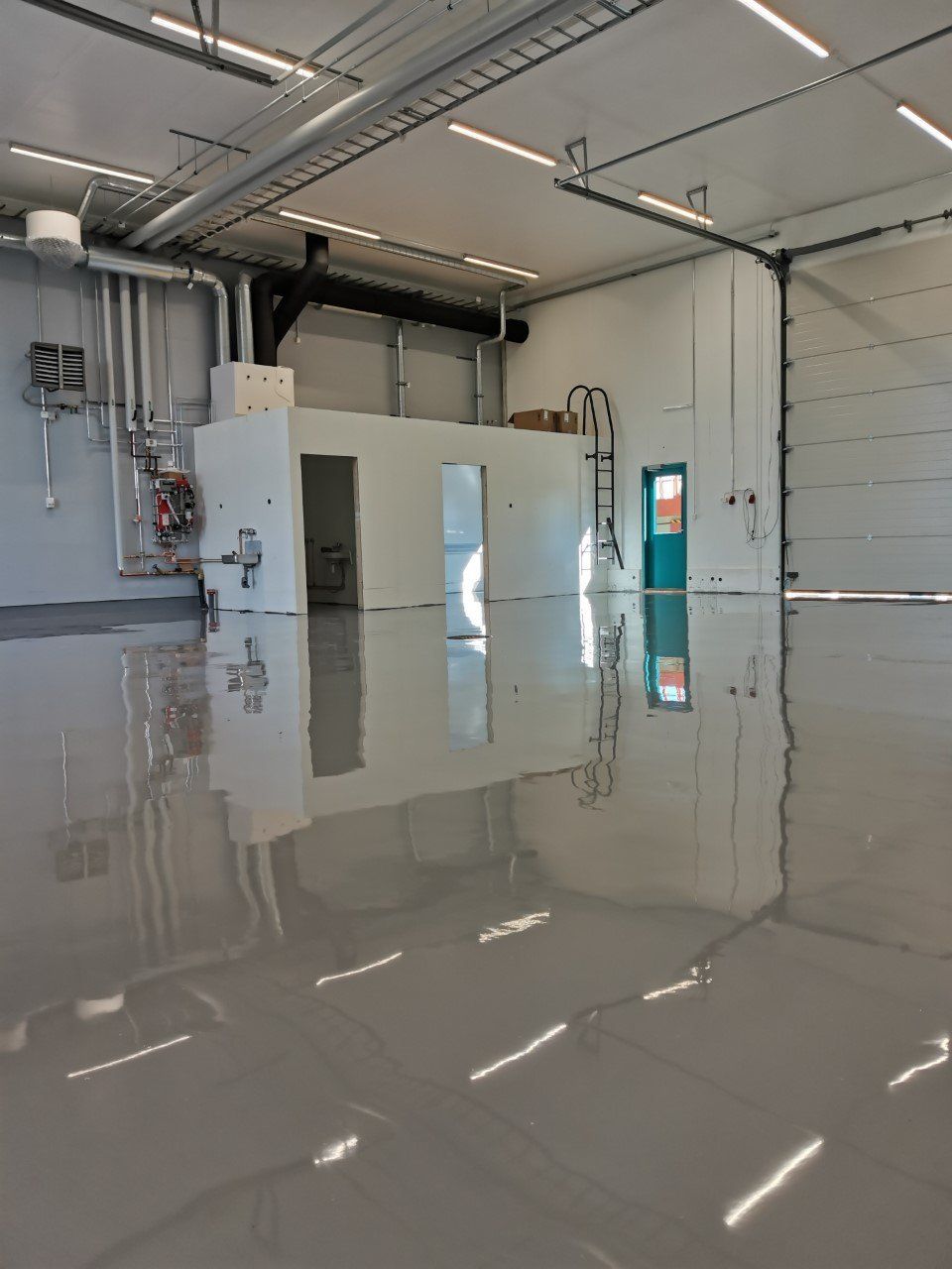 Tyhjä teollisuushuone, jossa kiiltävä, heijastava harmaa lattia; ilmanvaihto ja muut laitteet näkyvissä.