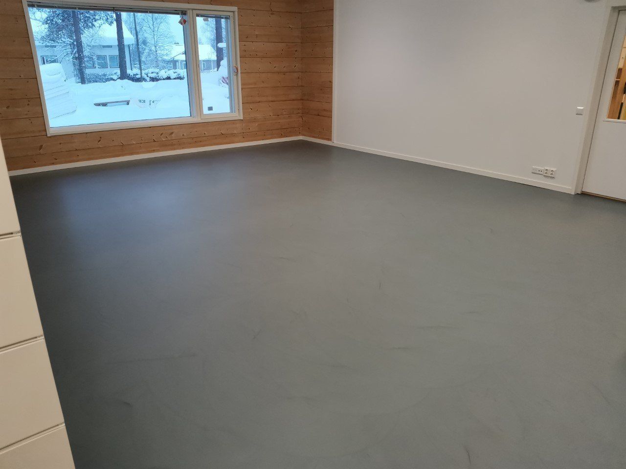 Tyhjä huone, jossa on harmaa lattia, valkoiset seinät ja puupaneloitu seinä. Ikkunasta näkyy luminen ulkonäkymä.