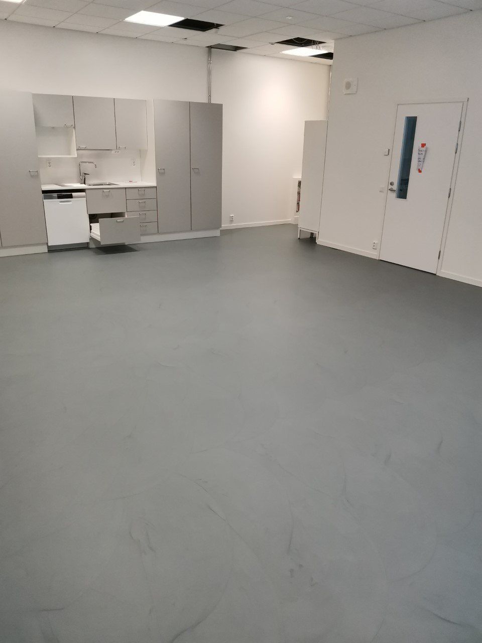 Tyhjä huone, jossa on harmaa lattia, valkoiset seinät ja kaapit sekä oviaukko.