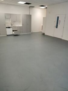 Tyhjä huone, jossa on harmaa lattia, valkoiset seinät ja pieni keittiötila kaapeilla.