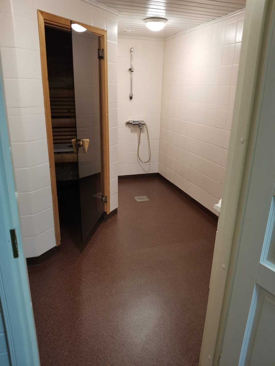 Suihkuhuone, jossa valkoiset laattaseinät, ruskeapilkkuinen lattia ja avoin saunan ovi.