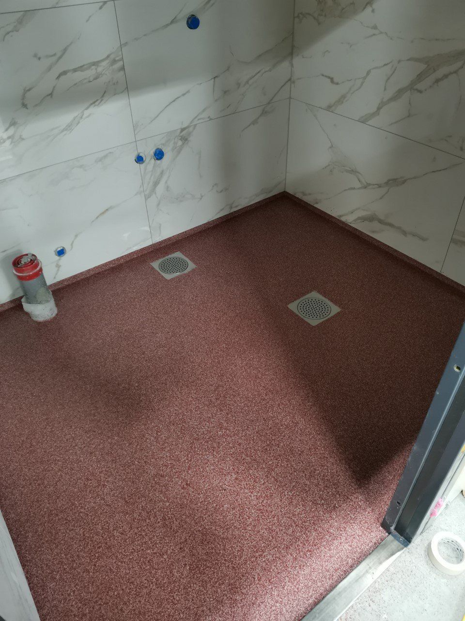 Suihkukaappi, jossa on punapilkkuinen lattia, valkoiset marmorijäljitelmälaatat ja kaksi viemäriä.