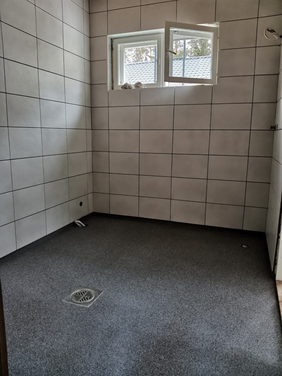 Pieni rakenteilla oleva kylpyhuone, jossa on harmaa lattia ja valkoiset laattaseinät. Takana on pieni ikkuna.