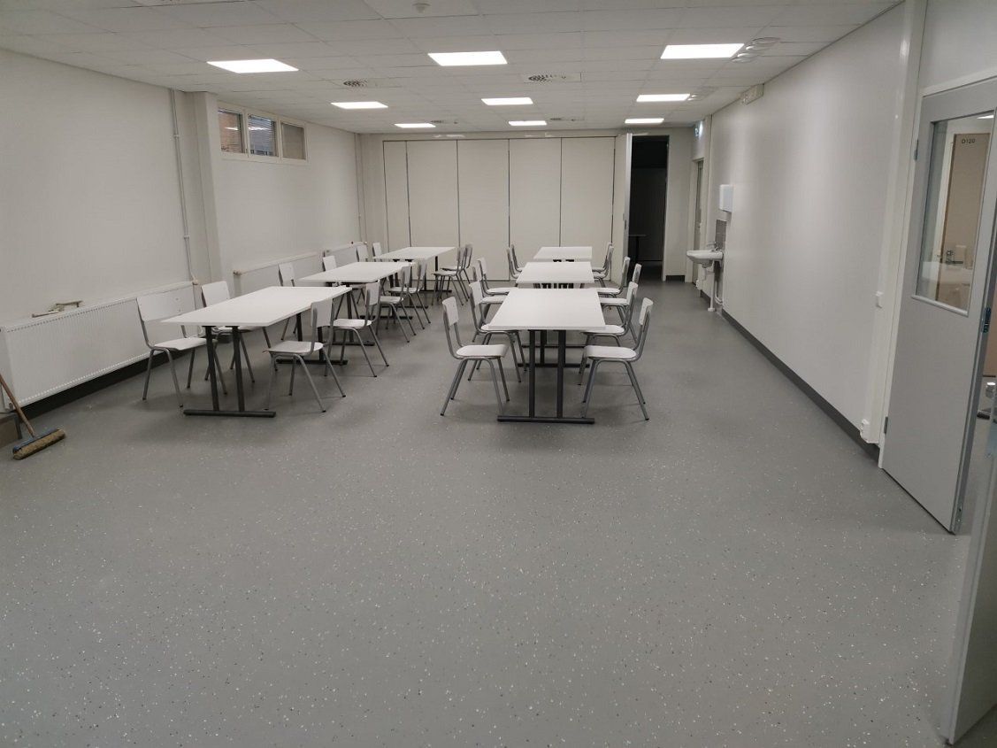 Tyhjä huone, jossa on pöytiä ja tuoleja, harmaa lattia, valkoiset seinät ja ovi oikealla.