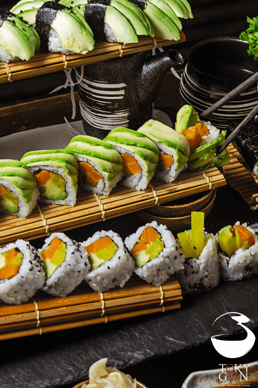Sushi-Rollen, angerichtet auf Bambusmatten, mit Avocado, Gurke und gelben Füllungen.