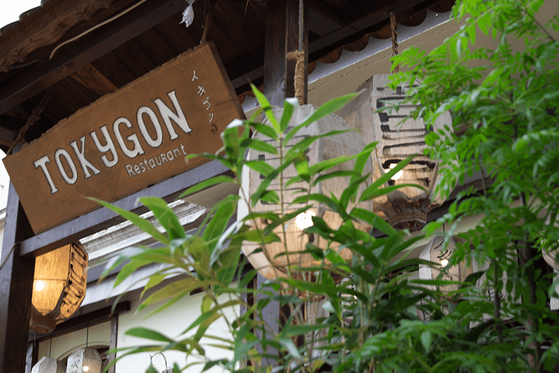 Schild für das Restaurant TOKYGON, braunes Holzschild mit weißer Schrift, umgeben von Grünpflanzen.