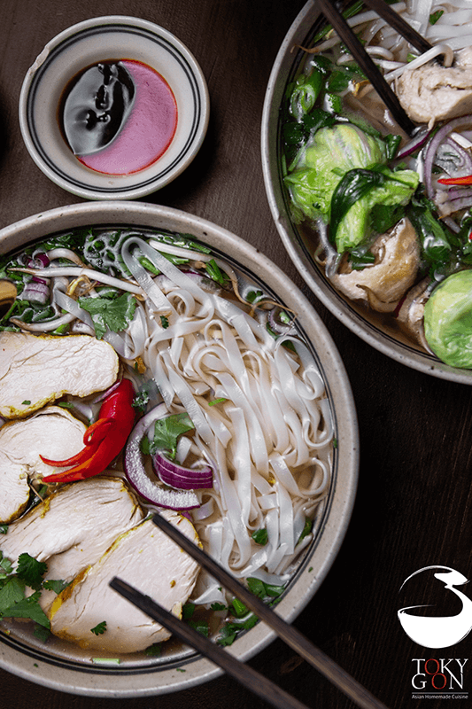 Schüsseln mit vietnamesischer Pho-Suppe, bestehend aus Hühnchen, Gemüse und Nudeln, serviert mit Sojasauce auf dunklem Untergrund.