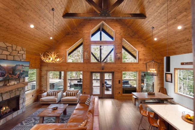 Spacious cabin interior with wooden walls, fireplace, leather sofa, and large windows with outdoor views.