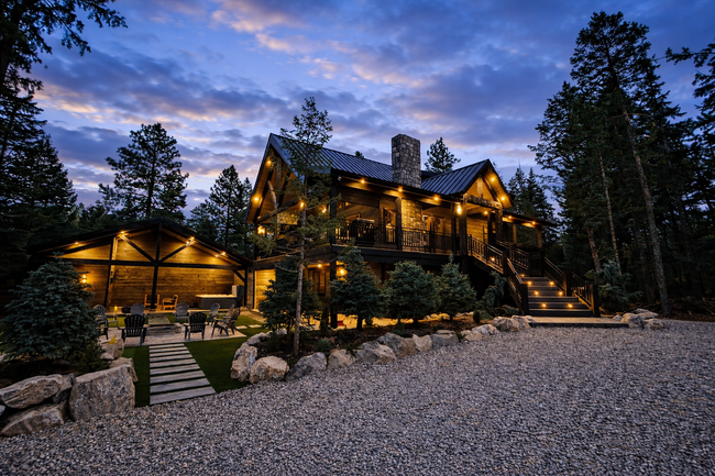 Rustic cabin illuminated at dusk, surrounded by trees. Stone exterior, gravel driveway, and outdoor seating.