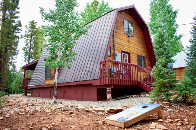 A rustic wooden A-frame cabin with a metal roof and deck, set among pine trees with a cornhole board in the foreground.