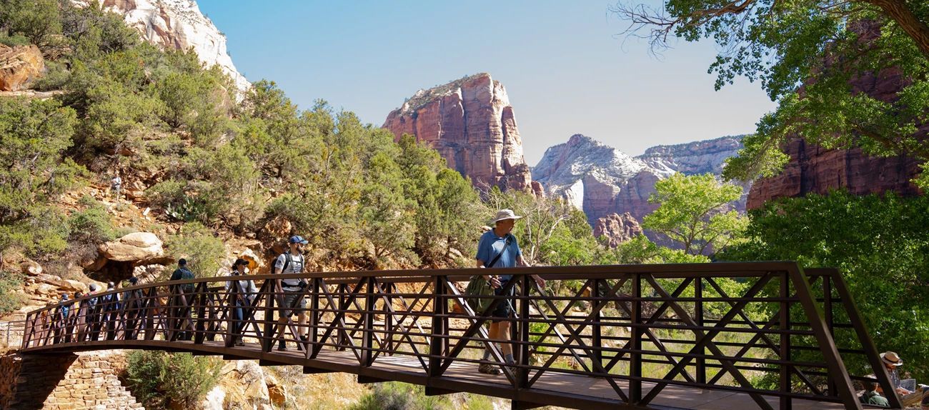 People walk across a wooden bridge in a canyon with lush greenery and mountains in the background.