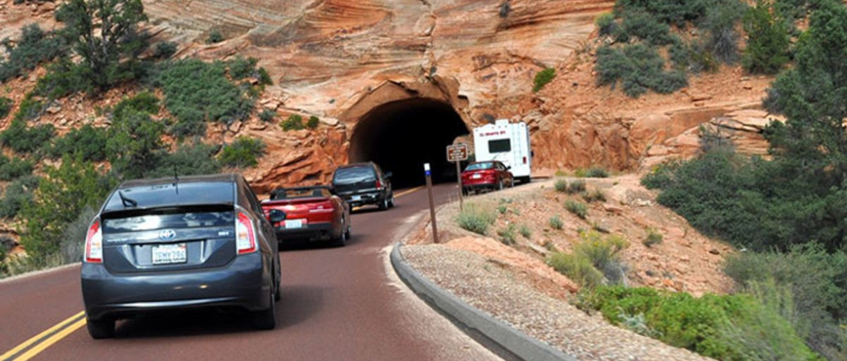 Cars driving on a road towards a tunnel carved in a rock formation; a camper sits near the entrance.
