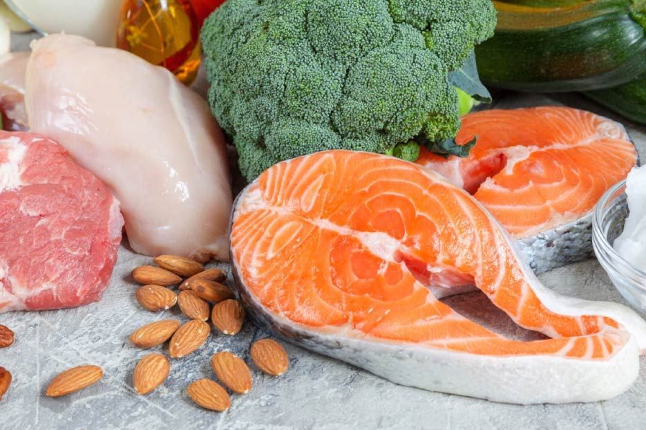 Una variedad de alimentos saludables que incluyen salmón, brócoli, carne, almendras y verduras en una mesa.