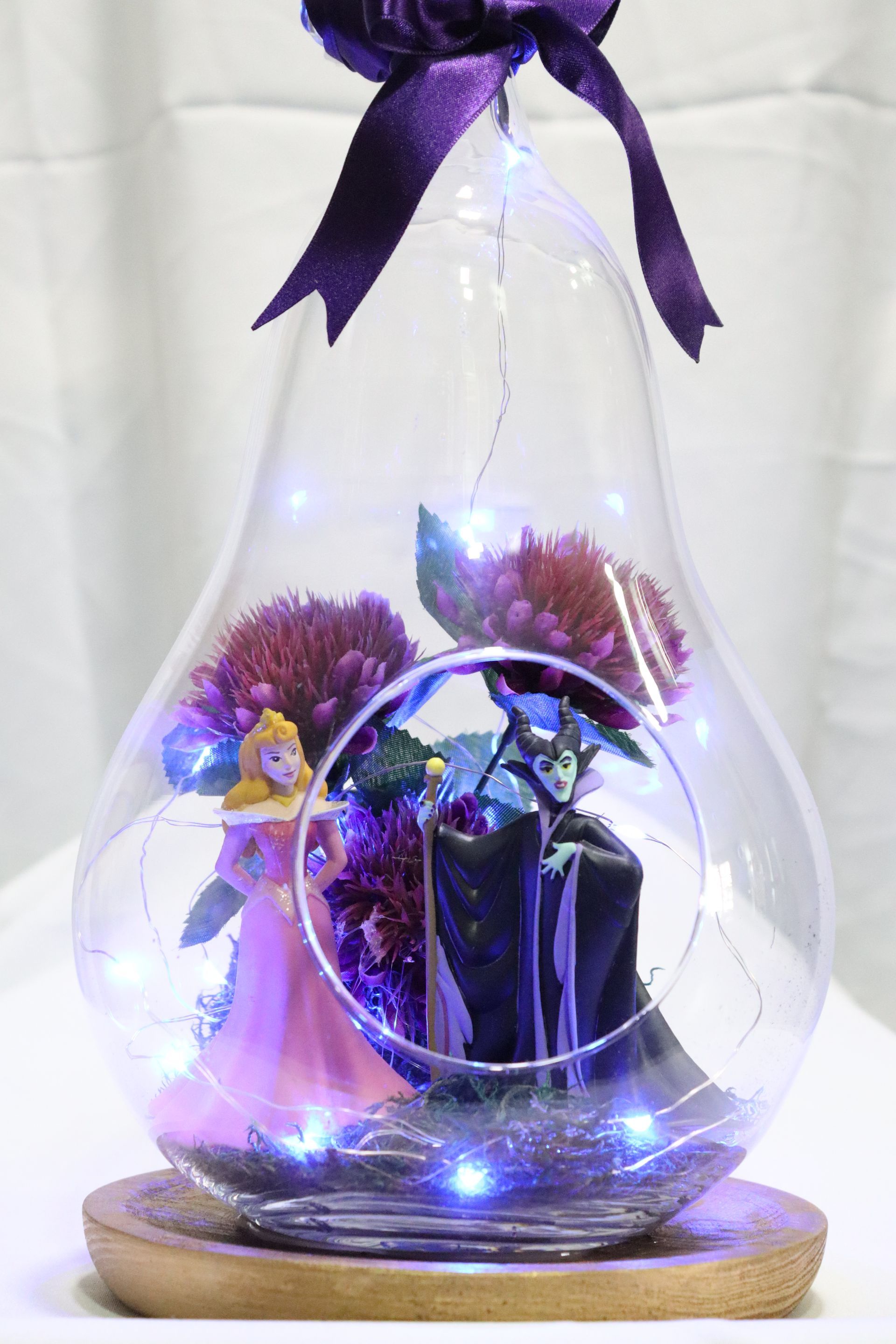 Figuras de La Bella Durmiente y Maléfica en un terrario de cristal con flores, luces y lazo morado.