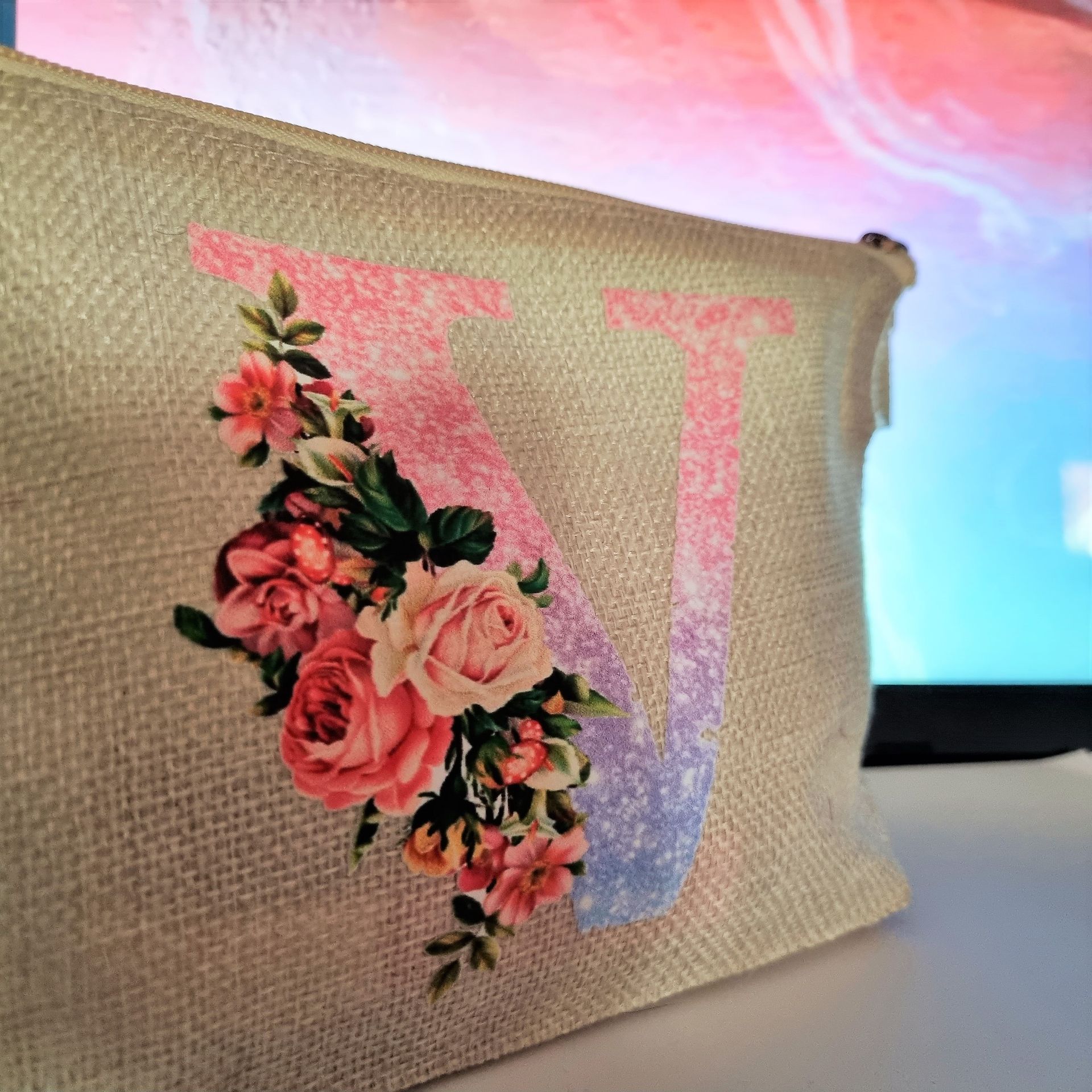 Bolsa de maquillaje beige con la letra “V” en degradado rosa y azul, decorada con ramo de flores rosas.