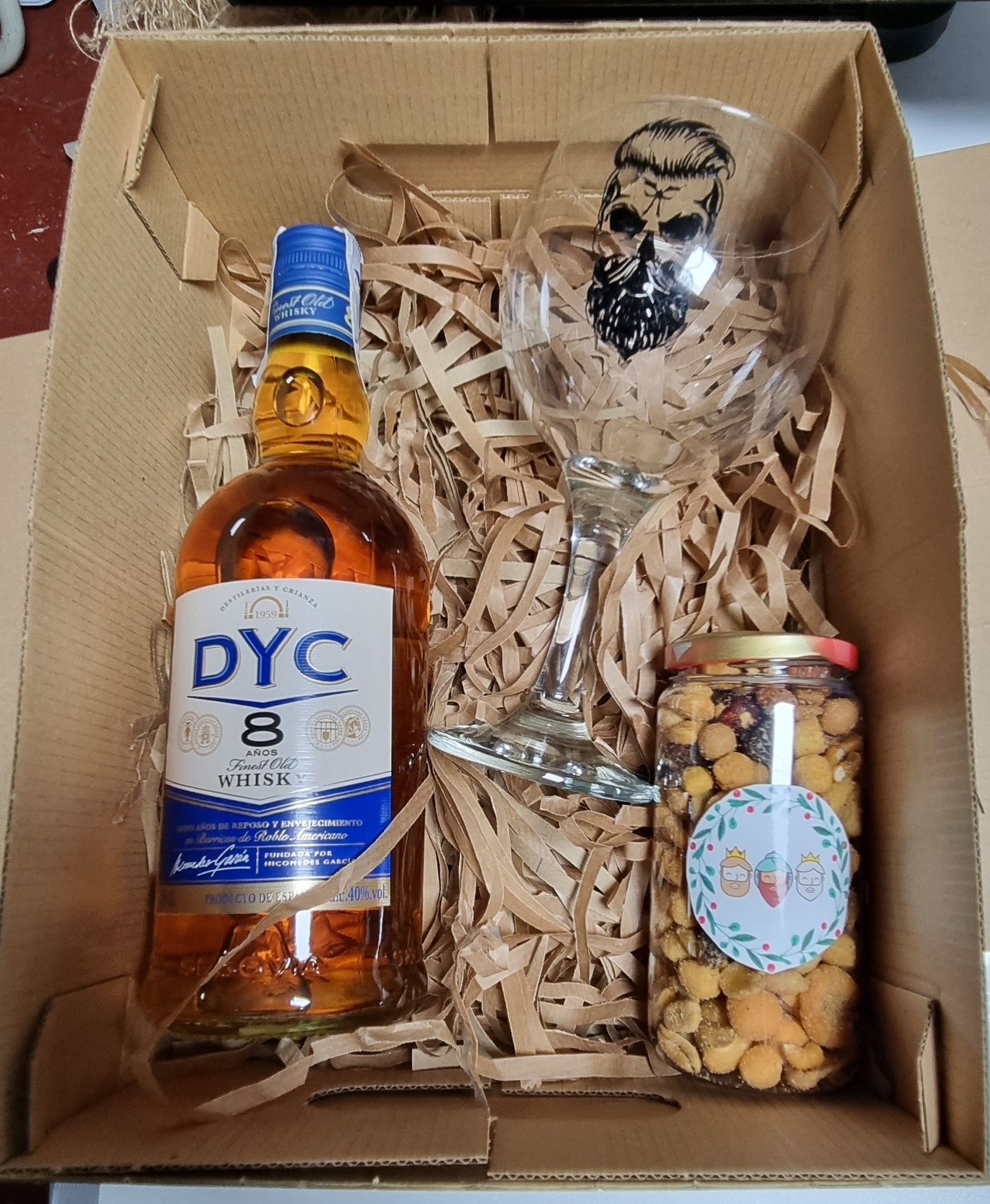 Caja de regalo: Botella de whisky DYC, un vaso y un tarro de snacks, todo ello envuelto en papel.