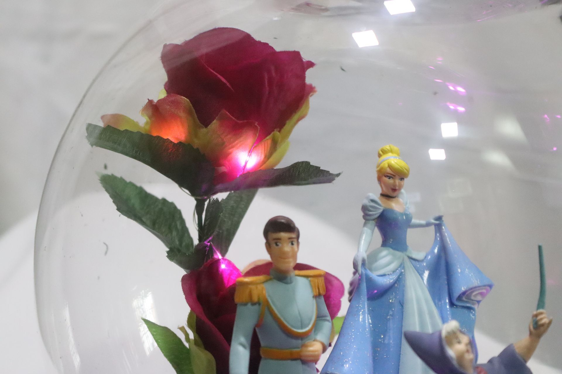 Figuras de Cenicienta y el Príncipe junto a una rosa bajo una cúpula de cristal con una flor iluminada.