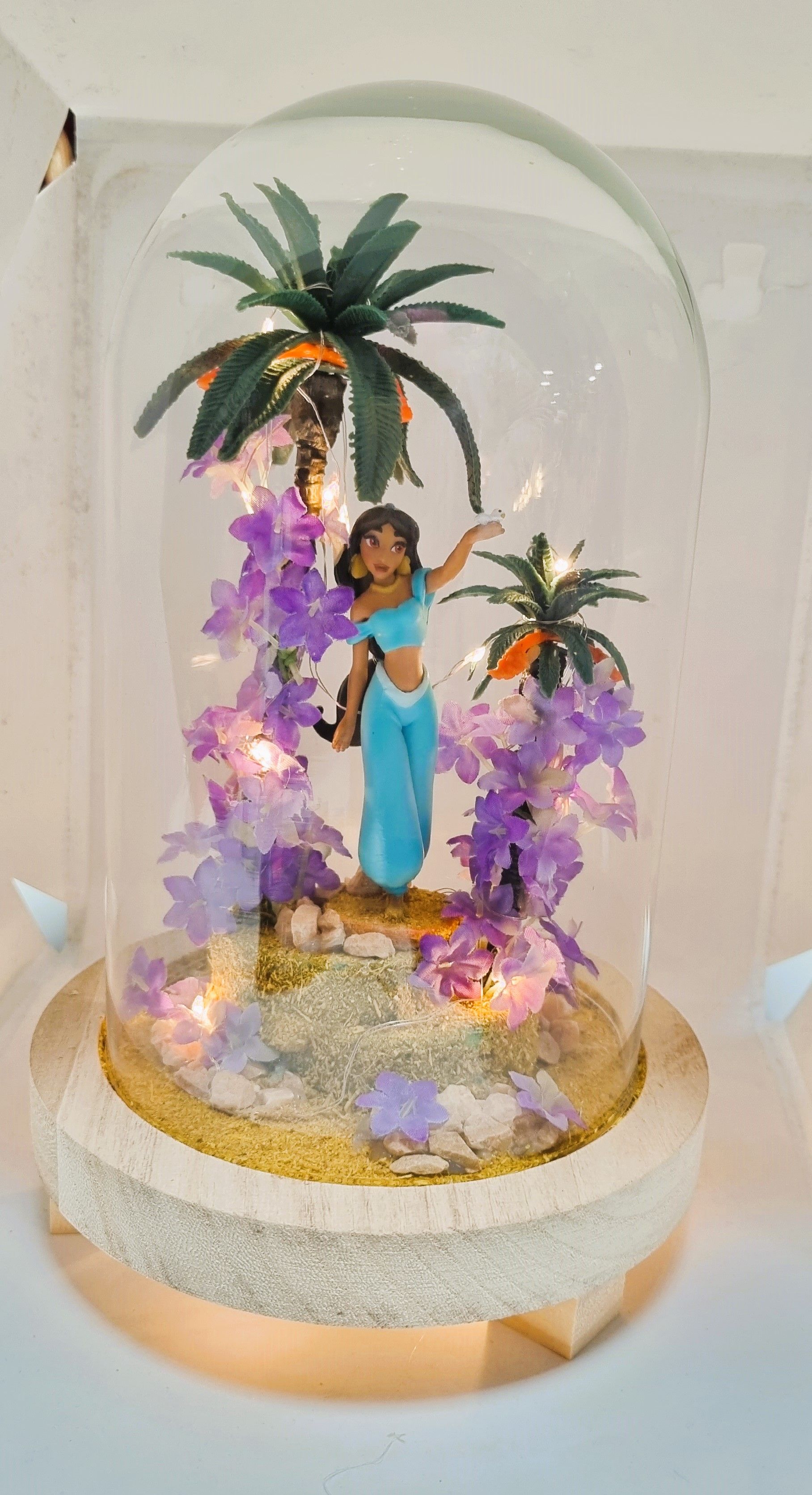 Figura de la princesa Jasmine en una cúpula de cristal, rodeada de flores, palmeras y luces.